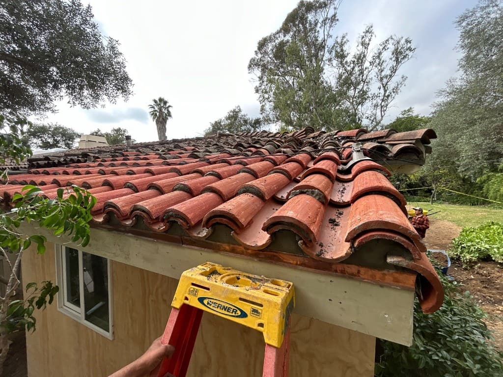 Clay tile roof repair on a Santa Barbara outbuilding with ladder and copper flashing at the eave